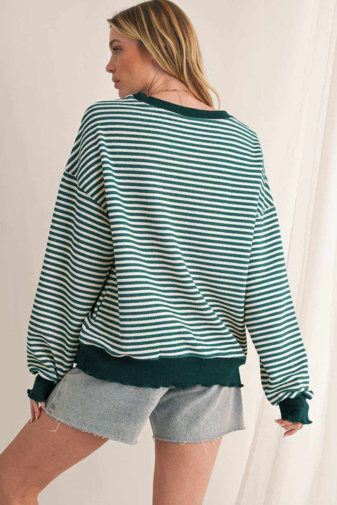 Woman wearing a green and white striped drop shoulder long sleeve top with dark green cuffs and hem paired with denim shorts.