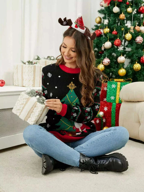 Woman wearing Christmas Tree Santa Round Neck Sweater sitting by decorated Christmas tree holding a gift box