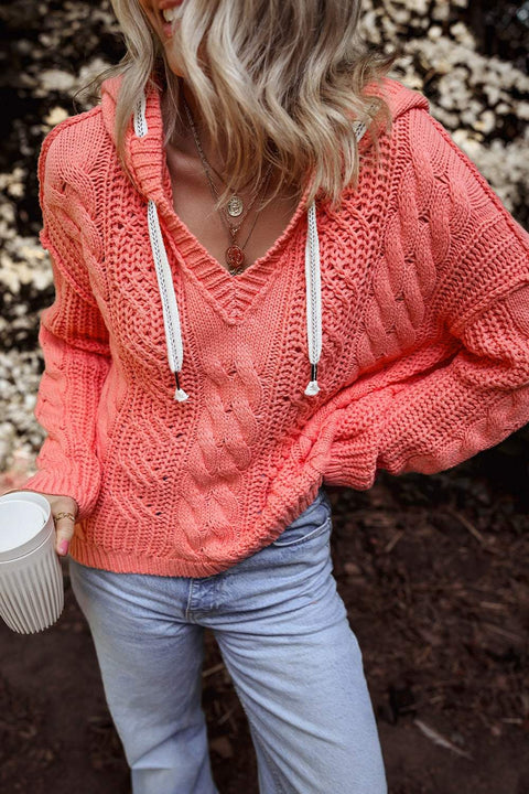 Cable-knit hooded sweater with drawstring in coral color, worn with jeans.