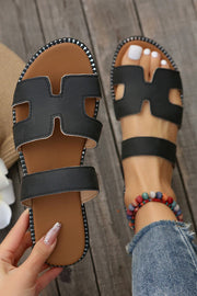 Black leather H band slides with cushioned footbed on wooden floor.
