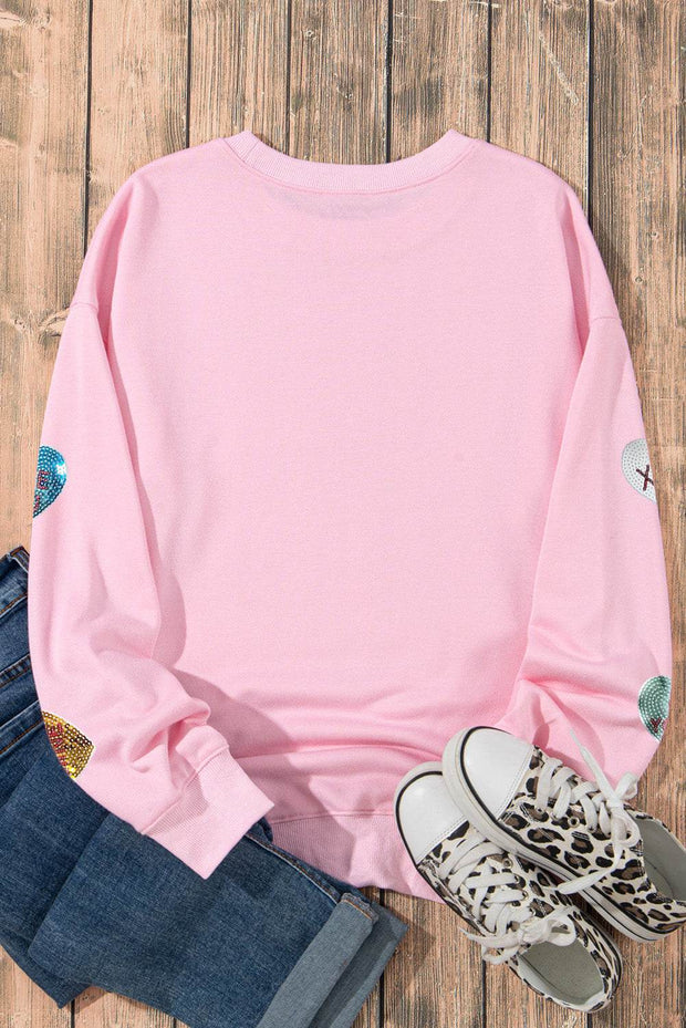 Pink graphic Valentines sweatshirt with sequined heart details, paired with jeans and leopard print sneakers on wooden background.