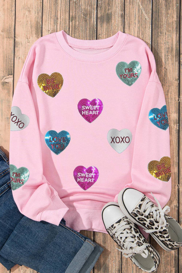 Pink graphic Valentines sweatshirt with sequin heart graphics and romantic lettering on a wooden background.