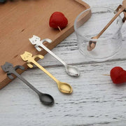 Stainless Steel Cat Teaspoons with whimsical cat-shaped handles in silver, gold, and black, on a wooden tray next to a glass cup and heart-shaped decor.