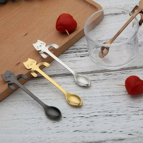 Stainless Steel Cat Teaspoons with whimsical cat-shaped handles in silver, gold, and black, on a wooden tray next to a glass cup and heart-shaped decor.